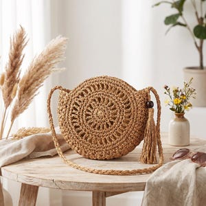 May include: A round, tan woven straw crossbody bag featuring an intricate circular pattern, a long braided shoulder strap, and a decorative tassel attached to the side with a dark brown wooden bead accent.