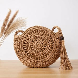 May include: A round, tan woven straw handbag featuring a circular open-work pattern on the front. The bag includes a matching woven strap and a decorative tassel accent attached to the side with a single dark brown wooden bead at the top.