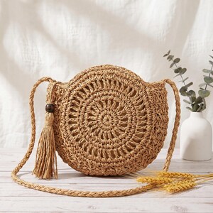 May include: A round, tan woven straw crossbody bag featuring a circular pattern, a long braided shoulder strap, and a decorative tassel accent attached to the side with a single dark brown wooden bead. The bag has a structured, circular shape.