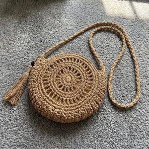 May include: A round, tan woven straw crossbody bag featuring a circular geometric pattern, a long braided shoulder strap, and a decorative tassel attached to the side with a small dark brown bead. The bag has a light beige fabric interior lining.