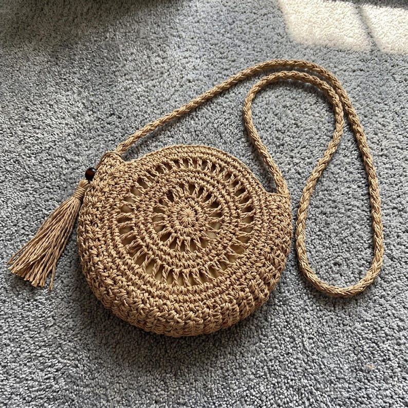 May include: A round, tan woven straw crossbody bag featuring a circular geometric pattern, a long braided shoulder strap, and a decorative tassel attached to the side with a small dark brown bead. The bag has a light beige fabric interior lining.
