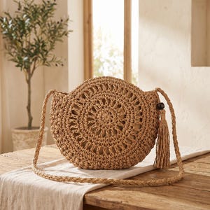 May include: A round, tan, woven straw crossbody bag featuring an intricate, open-work circular pattern on the front. The bag includes a long, braided shoulder strap and a single decorative tassel attached to the side with a dark brown wooden bead.