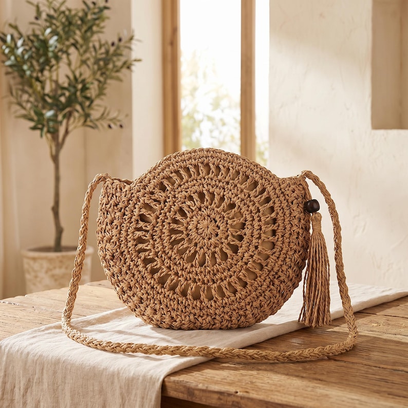 May include: A round, tan, woven straw crossbody bag featuring an intricate, open-work circular pattern on the front. The bag includes a long, braided shoulder strap and a single decorative tassel attached to the side with a dark brown wooden bead.