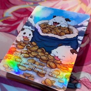 May include: A holographic trading card featuring cartoon characters and gold coins. The card depicts a white creature with a blue bag overflowing with gold coins. Text on the card reads "GOLD" and "KILL THIS REACTION ADD ABILITIES THAT ADD RESOURCES CANNOT BE REACTED TO."