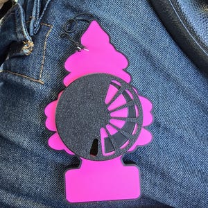 May include: A bright pink air freshener shaped like a stylised evergreen tree. The centre features a black circular overlay with a cut-out design resembling a fan or wheel with radiating spokes. A thin black elastic string is attached to the top.
