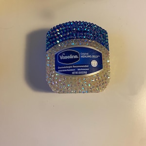 May include: A bedazzled jar of Vaseline Original Healing Jelly. The top rim is covered in blue rhinestones, while the body is covered in clear rhinestones. The label reads "Vaseline. Original Healing Jelly". The net weight is 1.75 oz.