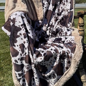 May include: A plush throw blanket featuring a brown and white cow print pattern on one side and a solid tan, textured sherpa fleece lining on the reverse. The blanket is draped over a wooden chair, showcasing its soft, fuzzy material and edges.