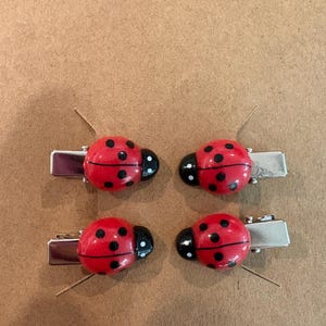 May include: Four hair clips featuring red ladybug charms with black spots and black heads. Each ladybug is attached to a silver-toned metal alligator clip. The ladybugs have a rounded, glossy finish and are arranged in a two-by-two grid pattern.