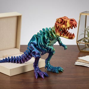 Puede incluir: Una colorida figura de esqueleto de Tyrannosaurus Rex impresa en 3D. El dinosaurio presenta un degradado de colores que va del morado al azul, verde y dorado. La figura está colocada sobre una superficie de madera, con una caja beige y objetos decorativos en el fondo.