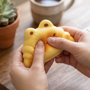 May include: A yellow, crescent-shaped squishy toy designed to resemble a croissant, featuring a soft, foam-like texture with small brown spots on the top surface. The object is being compressed between two hands, demonstrating its flexible material.