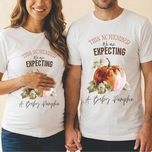 Puede incluir: Dos camisetas blancas de manga corta con un gráfico de calabaza en acuarela y hojas verdes. El texto dice: THIS NOVEMBER We are EXPECTING A Baby Pumpkin. El diseño está centrado en el pecho de cada camiseta con letras marrones y negras.
