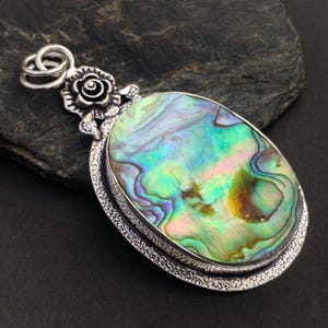 May include: An oval pendant featuring a large, iridescent abalone shell inlay with swirling green, blue, and pink tones. The shell is set in a textured silver-toned metal frame with a decorative rose-shaped bail at the top and a double ring loop.
