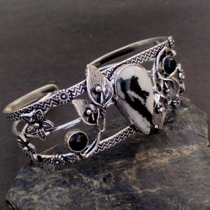 May include: A silver-toned cuff bracelet featuring a central teardrop-shaped white stone with black veining. The metal band is decorated with embossed zigzag patterns, small floral motifs, and two round, smooth black stones set on the sides.