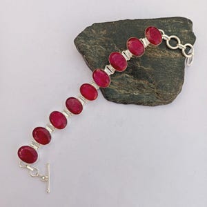 Könnte beinhalten: Ein Silberarmband mit zehn ovalen, tiefroten Rubinedelsteinen in polierten Metallfassungen. Das Armband verfügt über einen Knebelverschluss an einem Ende, wobei jeder Stein durch kleine Silberstege in einer linearen Anordnung verbunden ist.