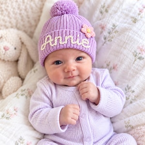 Puede incluir: Un gorro de punto acanalado de color morado claro con un pompón a juego en la parte superior, una pequeña aplicación de flor de ganchillo rosa y amarilla, y el nombre Annie bordado en cursiva blanca en el frente del dobladillo.