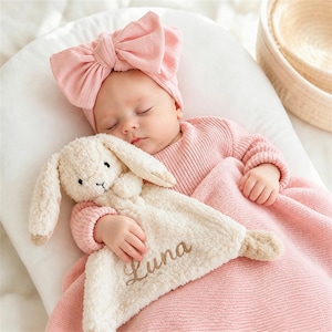 May include: A cream-colored plush bunny security blanket featuring the embroidered name Luna in brown script. The soft, textured fabric toy is held by a sleeping infant wearing a light pink ribbed long-sleeve shirt and a matching pink bow headband.