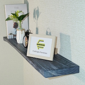 May include: A dark gray wooden floating shelf with a visible wood grain texture. On the shelf sits a white book titled The Kinfolk, a white geometric vase with artificial flowers, a black analog alarm clock, and a white frame with the text Craftopia Furniture.