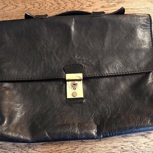May include: A dark brown leather briefcase featuring a rectangular flap closure and a central gold-toned metal push-lock clasp. The bag has a smooth, slightly textured surface with visible stitching along the edges and a top handle.