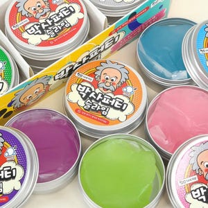 May include: A collection of round metal tins containing colorful slime, each featuring a cartoon scientist character on the lid with the Korean text 박사퍼티 슬라임. The visible slime colors include translucent blue, pink, purple, and bright green.