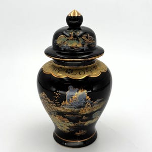 May include: A black ceramic ginger jar with a matching lid featuring a gold finial. The jar is decorated with gold and muted blue landscape illustrations of mountains, trees, and boats, with a decorative gold scalloped band around the neck.