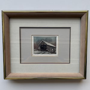 May include: Framed artwork depicting a snow-covered covered bridge. The artwork is in a square format, with a muted color palette of browns, grays, and whites. The frame is gold-toned with a light gray matting.