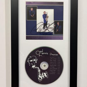 May include: A framed display featuring a Carl Ferris Promises CD and three photographs of a musician playing the saxophone. The CD label is purple with white text and a line drawing of a person. The photos are signed with a black marker.