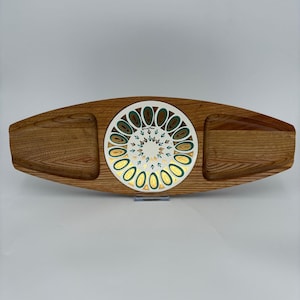 May include: An elongated wooden serving tray with a central circular ceramic inlay. The wood features a prominent grain pattern. The white ceramic center displays a radial pattern of teal and gold teardrop shapes and small leaf-like motifs.