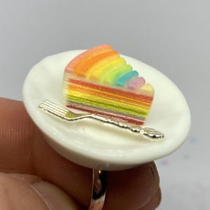 Può includere: Anello con una fetta di torta arcobaleno in miniatura su un piatto bianco con una forchetta d'argento. La torta presenta strati di colori vivaci: rosso, arancione, giallo, verde, blu e viola. L'anello è argentato.
