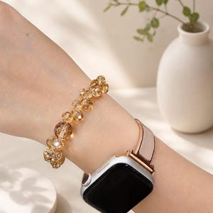 Puede incluir: Una pulsera de cuentas doradas y un reloj inteligente con caja plateada y correa rosa claro se llevan en la muñeca. La pulsera presenta un diseño floral con múltiples cuentas transparentes y color ámbar. El reloj inteligente tiene una pantalla negra.