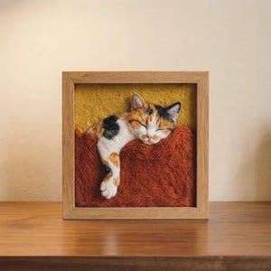 May include: A square wooden frame containing a needle-felted calico cat sleeping in a rust-colored blanket against a mustard yellow background. The cat has white, orange, and black fur, with one paw extended over the edge of the textured blanket.