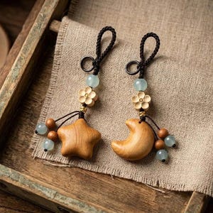 Puede incluir: Dos adornos colgantes con una estrella de madera y una luna creciente de madera. Cada pieza incluye una flor de madera tallada, una cuenta azul claro y lazos de cordón marrón oscuro. Pequeñas cuentas marrones y azul claro decoran la parte inferior.