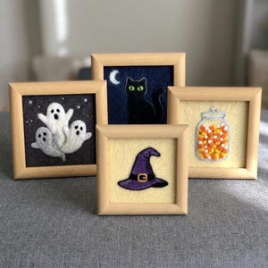 Puede incluir: Cuatro marcos de madera cuadrados con arte de fieltro de aguja. Uno muestra tres fantasmas blancos sobre fondo gris oscuro, otro un gato negro con una luna creciente, uno un sombrero de bruja morado y el último un frasco con dulces.