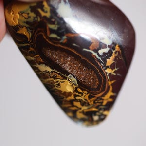 May include: A polished boulder opal gemstone featuring a dark brown base with intricate, swirling patterns of cream, yellow, and metallic gold. The centre displays a textured, granular cavity surrounded by deep brown and black concentric bands.