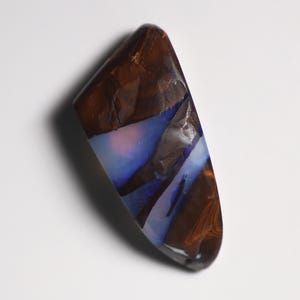 May include: A polished boulder opal gemstone with a dark brown ironstone matrix featuring irregular, fractured patterns. The stone displays vibrant flashes of iridescent blue and soft pink light play across its smooth, glossy surface.