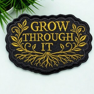 May include: An oval-shaped black embroidered patch with a scalloped edge. The patch features the gold-colored text "GROW THROUGH IT" centered above a detailed illustration of tree roots and leafy branches framing the words on both sides.