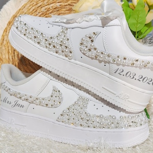 May include: A pair of white sneakers featuring white pearl and clear crystal embellishments on the side panels and swoosh logos. The shoes include white satin ribbon laces and custom text printed on the heels reading Mrs Jan and 12.03.2024.