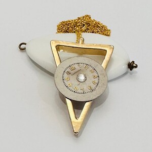 May include: A gold triangle pendant necklace with a white porcelain oval charm and a small clock face with a rhinestone in the center. The clock face reads "Corum" and has Roman numeral markings.