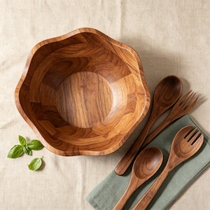 Puede incluir: Un cuenco de madera grande con bordes festoneados y un patrón de veta visible, acompañado de dos cucharas y dos tenedores de madera. Tres hojas de albahaca verde fresca se sitúan a la izquierda del cuenco, y los utensilios descansan sobre un paño verde.