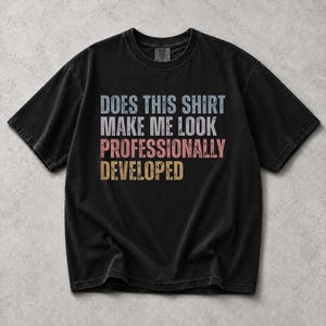 May include: A black short-sleeve t-shirt with a distressed graphic print on the chest. The text reads: DOES THIS SHIRT MAKE ME LOOK PROFESSIONALLY DEVELOPED. The lettering is in light blue, white, salmon pink, and tan, featuring a vintage style.