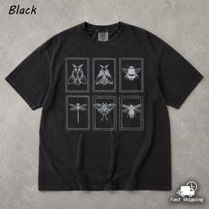 Może przedstawiać: Czarny t-shirt z krótkim rękawem z białym nadrukiem graficznym przedstawiającym sześć prostokątnych ramek w stylu znaczka pocztowego. Każda ramka zawiera szczegółowy rysunek liniowy innego owada, w tym różnych ciem, pszczoły i ważki.