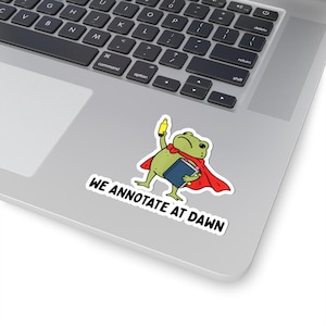 May include: A die-cut sticker featuring a green frog wearing a red cape, holding a yellow highlighter and a blue book. The frog has a serious expression. Below the frog, the text reads WE ANNOTATE AT DAWN in bold, black, capital letters.