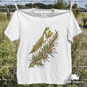 Puede incluir: Una camiseta blanca de manga corta con una ilustración gráfica detallada de una mantis religiosa verde posada sobre una rama de romero con pequeñas flores rosas. La camiseta tiene cuello redondo y una etiqueta visible en el cuello.
