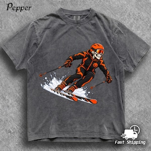 Op de afbeelding: Een antracietgrijs t-shirt met een afbeelding van een skelet in een oranje en zwart skipak, helm en skibril tijdens het skiën. Het skelet heeft een klein pompoenicoontje op de borst en er is witte sneeuw te zien.