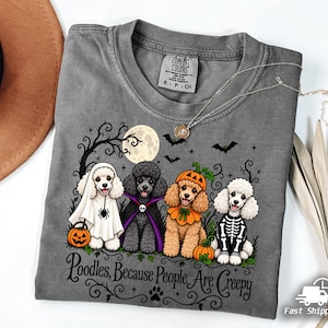 May include: A grey t-shirt featuring a graphic of four poodles in Halloween costumes, including a ghost, a vampire, a pumpkin, and a skeleton. The text reads, Poodles, Because People Are Creepy. The shirt has a crew neck and a Comfort Colors brand label.