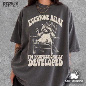 May include: A charcoal gray short-sleeve t-shirt featuring a graphic of a raccoon sitting in a chair. The text reads EVERYONE RELAX I'M PROFESSIONALLY DEVELOPED. The shirt has a crew neckline and a relaxed fit, with a gold chain necklace worn over the collar.