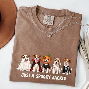 Könnte beinhalten: Ein braunes T-Shirt mit einer Grafik von fünf Jack Russell Terriern in Halloween-Kostümen, darunter ein Geist, ein Vampir, ein Kürbis, eine Mumie und ein Skelett. Der Text unter den Hunden lautet: "JUST A SPOOKY JACKIE."