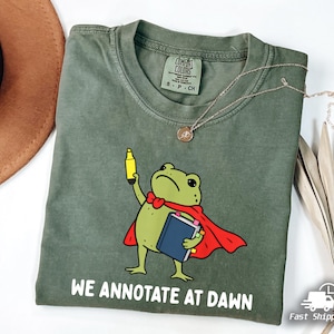May include: An olive green t-shirt featuring a graphic of a green frog wearing a red cape, holding a yellow highlighter and a blue book. The text "WE ANNOTATE AT DAWN" is printed in white below the frog. The shirt has a Comfort Colors brand label.