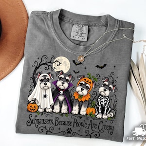 Op de afbeelding: Een grijs t-shirt met een afbeelding van vier Schnauzer-honden in Halloween-kostuums, waaronder een geest, een vampier, een pompoen en een skelet. De tekst luidt, Schnauzers, Because People Are Creepy, met een maan en vleermuizen.