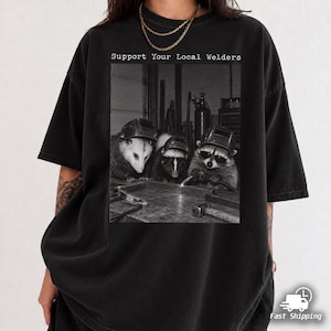 Op de afbeelding: Een zwart t-shirt met een grafische print van een opossum, een stinkdier en een wasbeer die lashelmen dragen en aan een werkbank werken. De tekst "Support Your Local Welders" staat in het wit boven de zwart-wit afbeelding gedrukt.