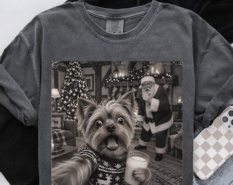 May include: A charcoal grey crew neck t-shirt featuring a black and white graphic print of a Yorkshire Terrier wearing a patterned sweater and holding a glass of milk, with Santa Claus standing in the background near a Christmas tree.